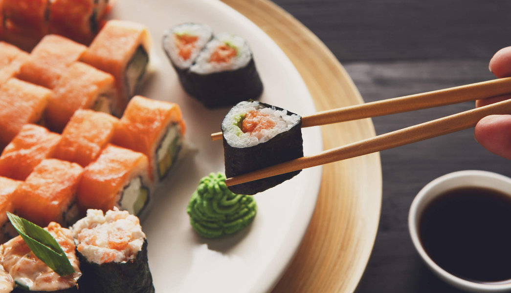 hashi segurando um sushi maki e ao fundo um prato com peças de comida japonesa
